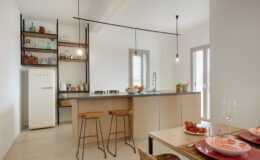 Modern kitchen interior featuring a sleek design with a white SMEG refrigerator, wooden bar stools, and a stylish countertop. The open shelving displays various kitchenware, while natural light streams in through large windows, enhancing the inviting atmosphere. A dining table is set with decorative plates and fresh tomatoes, creating a warm and functional space.