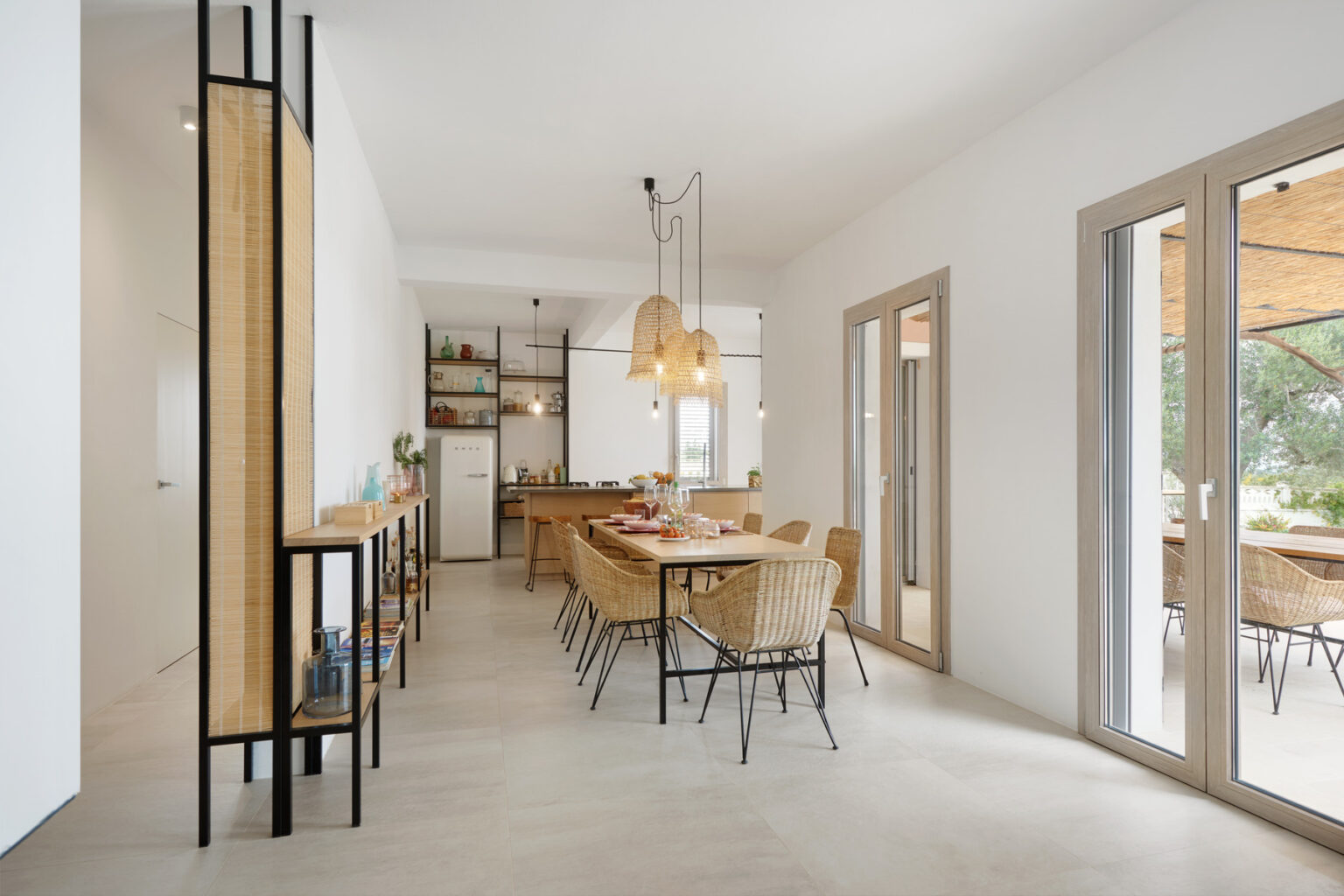 Modern dining area featuring a wooden table surrounded by stylish wicker chairs, complemented by pendant lighting and a sleek kitchen in the background. Large windows allow natural light to illuminate the space, creating an inviting atmosphere. | Only on SopranoVillas