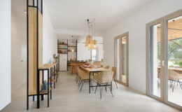 Modern dining area featuring a wooden table surrounded by stylish wicker chairs, complemented by pendant lighting and a sleek kitchen in the background. Large windows allow natural light to illuminate the space, creating an inviting atmosphere.