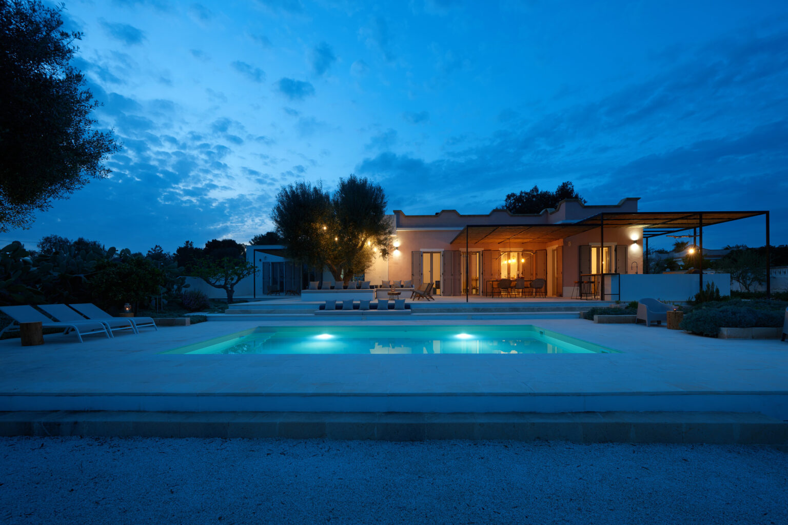Modern villa with illuminated swimming pool at dusk, surrounded by lush greenery and outdoor seating. Ideal for luxury vacation rentals or serene getaways. | Only on SopranoVillas