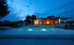 Modern villa with illuminated swimming pool at dusk, surrounded by lush greenery and outdoor seating. Ideal for luxury vacation rentals or serene getaways.