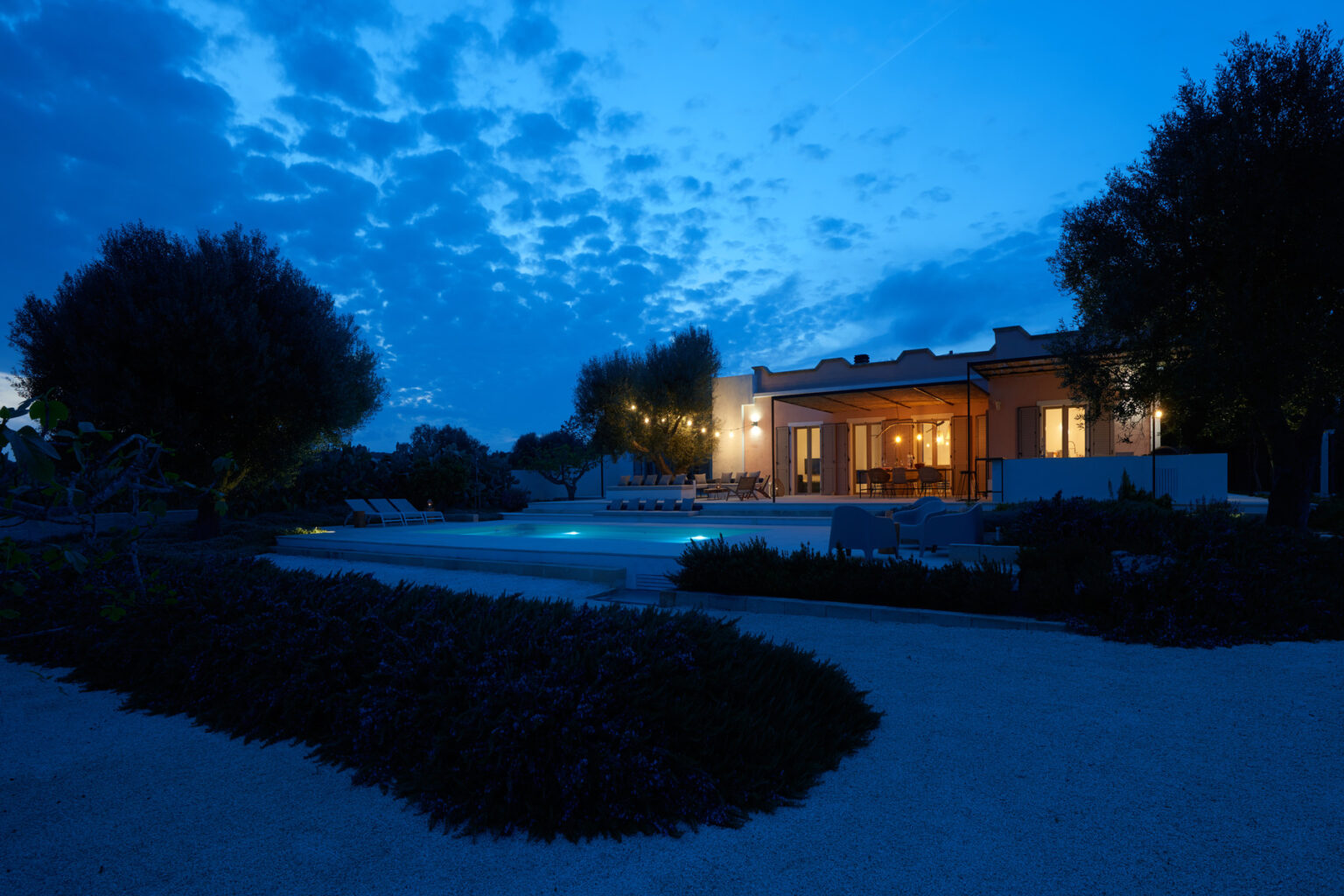 Modern house exterior at dusk with illuminated windows, surrounded by landscaped gardens and a tranquil pool area. | Only on SopranoVillas