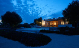 Modern house exterior at dusk with illuminated windows, surrounded by landscaped gardens and a tranquil pool area.