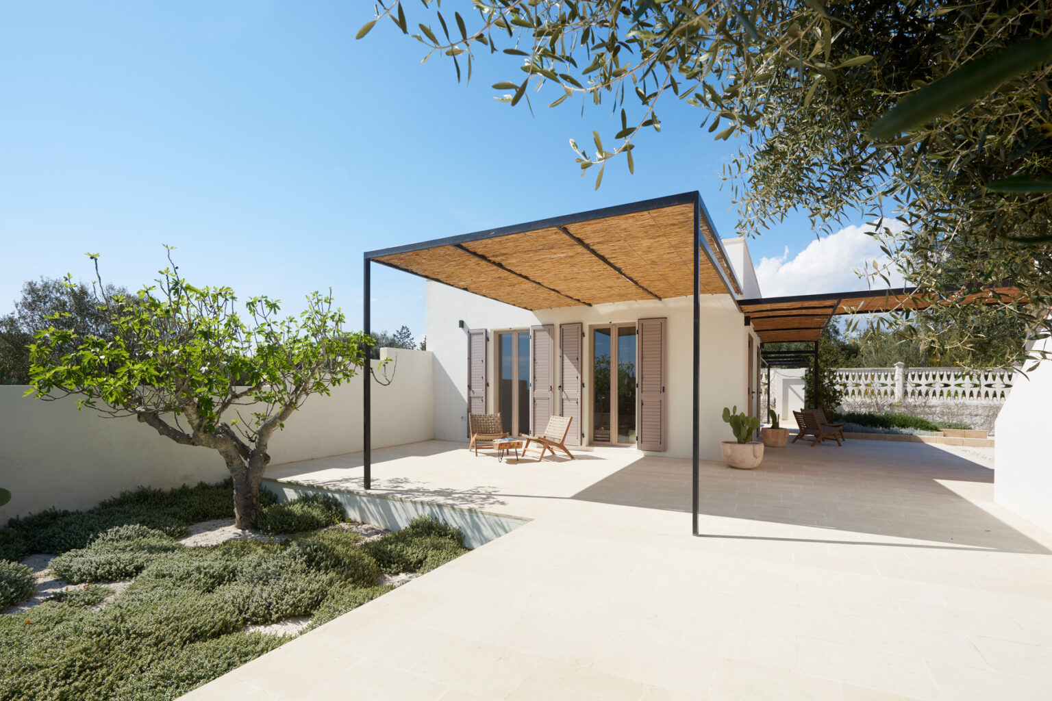 Modern minimalist house with a wooden pergola, surrounded by landscaped gardens and olive trees, featuring large glass doors and a serene outdoor seating area. | Only on SopranoVillas
