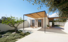 Modern minimalist house with a wooden pergola, surrounded by landscaped gardens and olive trees, featuring large glass doors and a serene outdoor seating area.