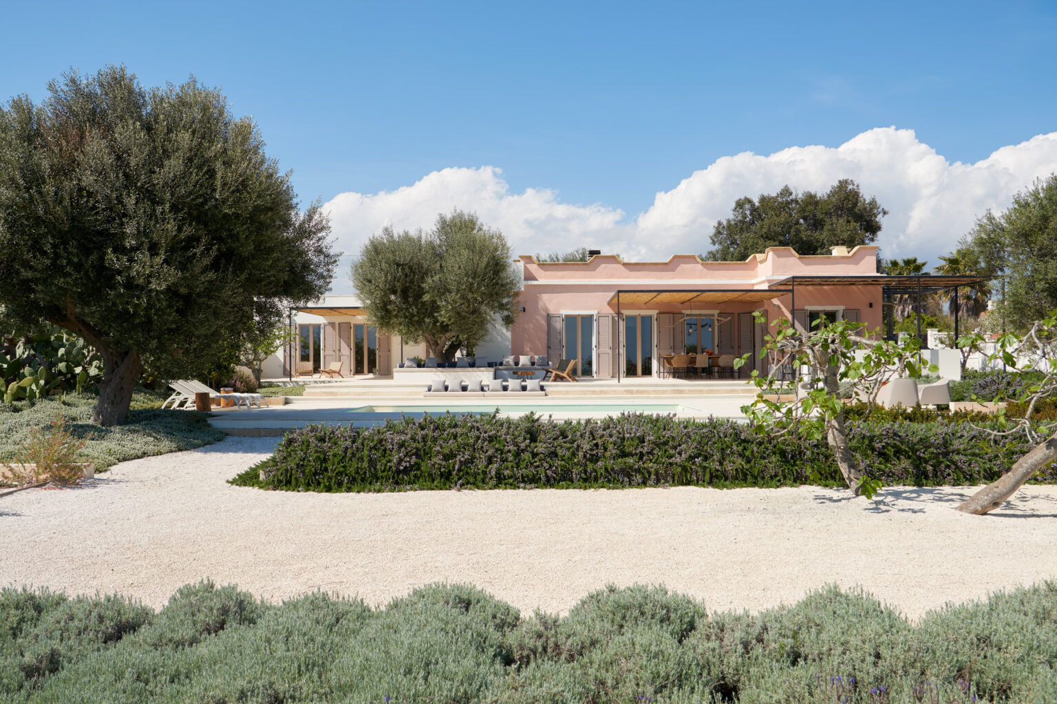 Modern villa with a pool surrounded by landscaped gardens, featuring olive trees and a sandy pathway, under a clear blue sky. Ideal for relaxation and outdoor living. | Only on SopranoVillas
