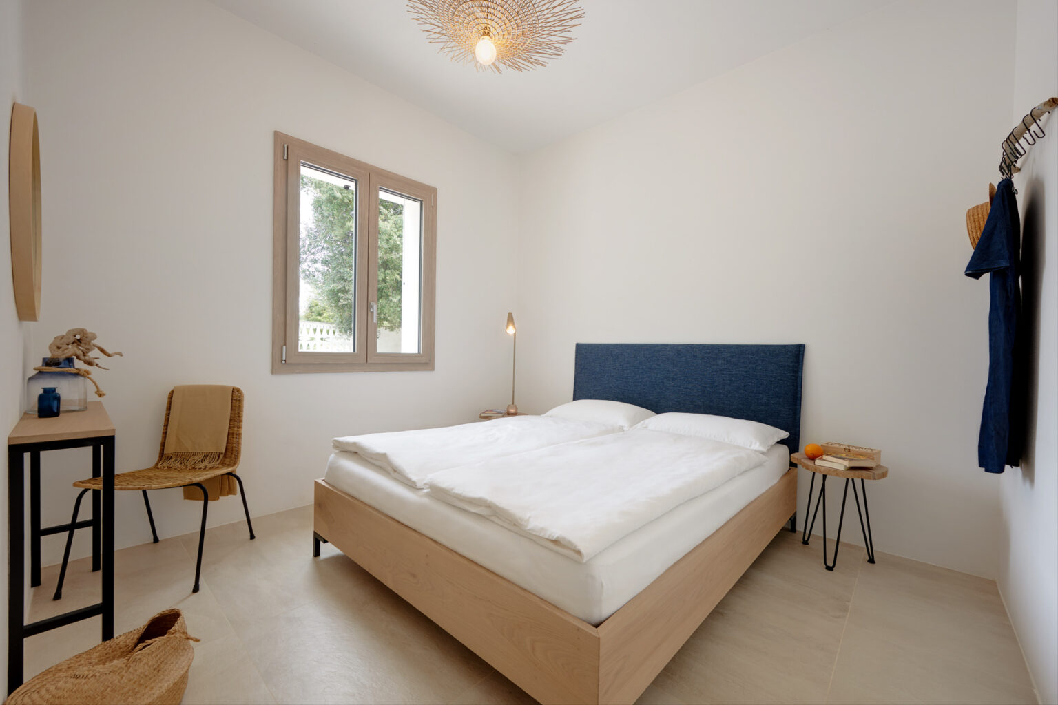 Modern bedroom interior featuring a double bed with a blue upholstered headboard, minimalistic decor, a wooden chair, and natural light coming through two windows. The room has a clean, neutral color palette and stylish lighting, creating a serene atmosphere. | Only on SopranoVillas