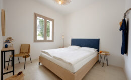 Modern bedroom interior featuring a double bed with a blue upholstered headboard, minimalistic decor, a wooden chair, and natural light coming through two windows. The room has a clean, neutral color palette and stylish lighting, creating a serene atmosphere.