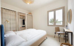 Modern bedroom with a double bed, natural light from a window, minimalist decor, a wooden chair, and a small desk, featuring a wardrobe with open shelving.