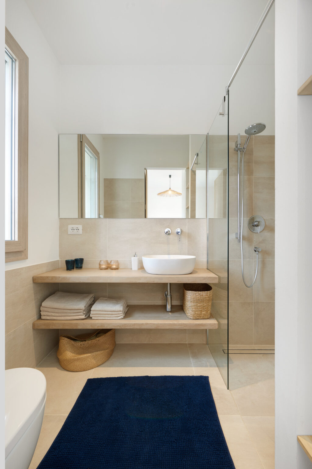 Modern bathroom interior featuring a wooden shelf with neatly stacked towels, a minimalist white sink, and a glass shower enclosure. Natural light illuminates the space through a window, enhancing the clean and contemporary design. | Only on SopranoVillas