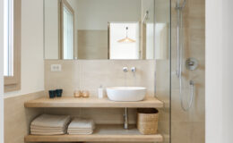 Modern bathroom interior featuring a wooden shelf with neatly stacked towels, a minimalist white sink, and a glass shower enclosure. Natural light illuminates the space through a window, enhancing the clean and contemporary design.