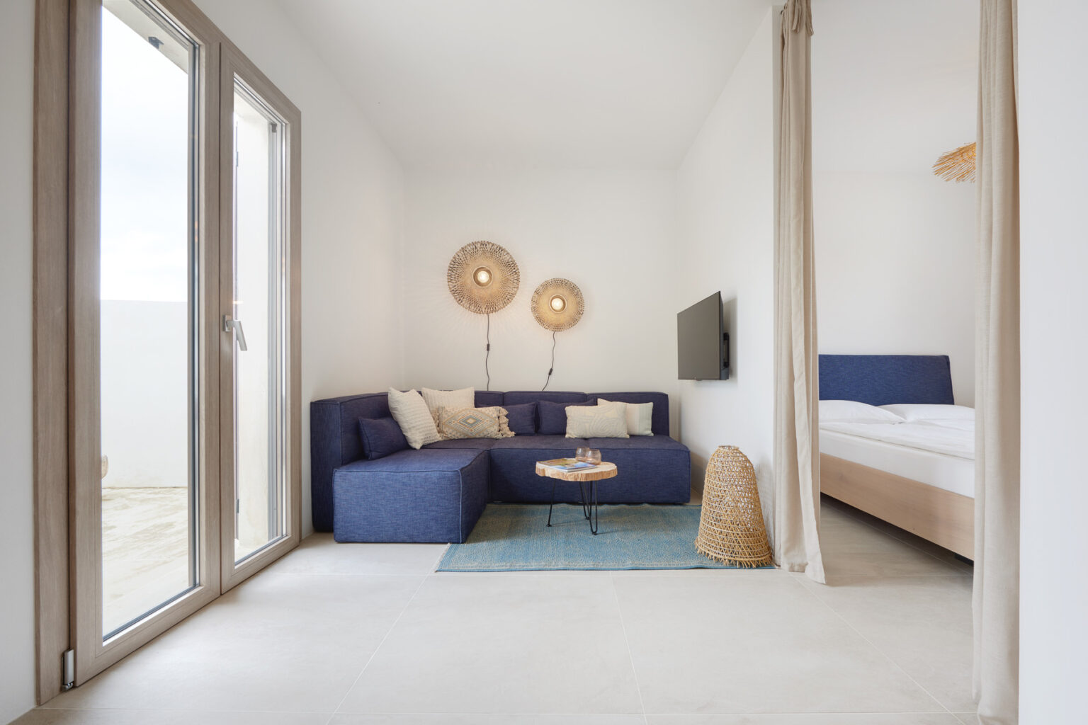 Modern living room with a blue sectional sofa, decorative wall lights, and a small coffee table, leading to a bedroom area with a light-colored bed. Large windows provide natural light in a bright, minimalist space. | Only on SopranoVillas