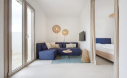 Modern living room with a blue sectional sofa, decorative wall lights, and a small coffee table, leading to a bedroom area with a light-colored bed. Large windows provide natural light in a bright, minimalist space.