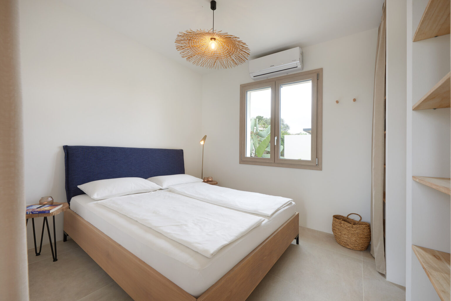 Cozy and modern bedroom featuring a double bed with white linens, a stylish blue headboard, natural light from a window, and minimalistic decor including a pendant light and wooden shelving. | Only on SopranoVillas