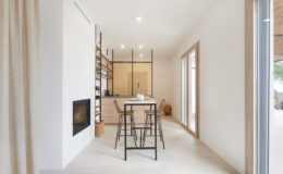 Modern minimalist kitchen and dining area featuring a sleek design with a small table and chairs, warm wood accents, and large windows allowing natural light to illuminate the space.