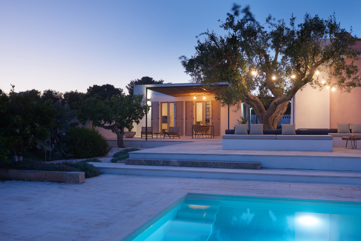 Modern villa with a swimming pool, surrounded by lush greenery, illuminated by soft evening lights, showcasing outdoor living space and contemporary architecture. | Only on SopranoVillas