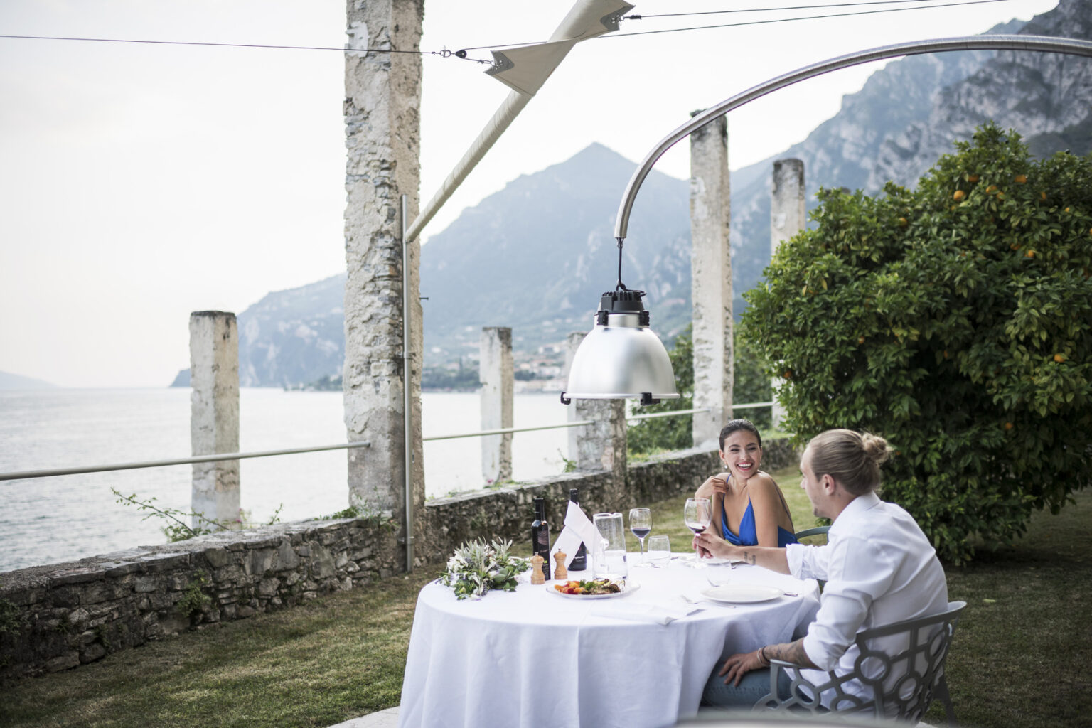 Couple enjoying a romantic dinner by the lake, with a scenic view of mountains and greenery, under a stylish lamp at a beautifully set table. | Only on SopranoVillas