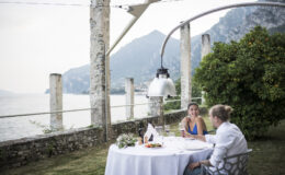 Couple enjoying a romantic dinner by the lake, with a scenic view of mountains and greenery, under a stylish lamp at a beautifully set table.