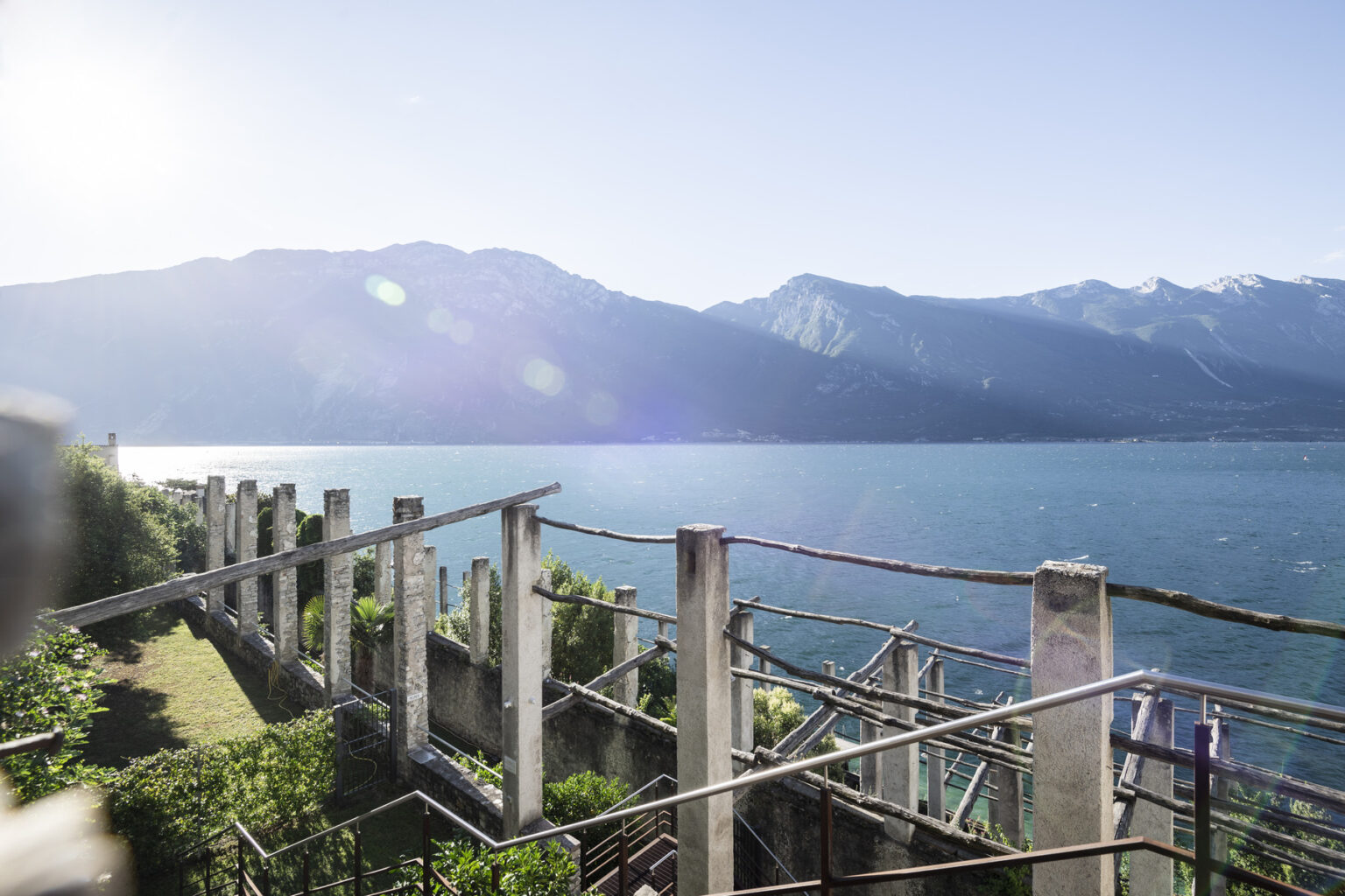 Scenic view of a lake surrounded by mountains, featuring a rustic terrace with plants and sunlight reflecting off the water. | Only on SopranoVillas
