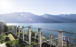 Scenic view of a lake surrounded by mountains, featuring a rustic terrace with plants and sunlight reflecting off the water.