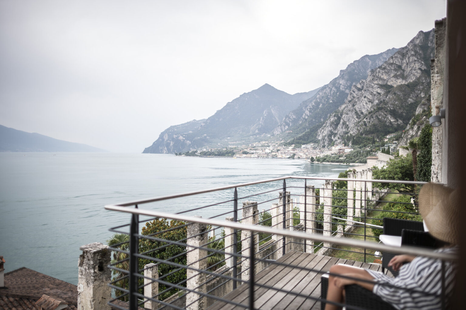 A serene view of a person relaxing on a balcony overlooking a lake surrounded by mountains, with a cloudy sky in the background. The scene captures the tranquility of nature and outdoor living. | Only on SopranoVillas