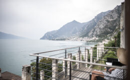 A serene view of a person relaxing on a balcony overlooking a lake surrounded by mountains, with a cloudy sky in the background. The scene captures the tranquility of nature and outdoor living.