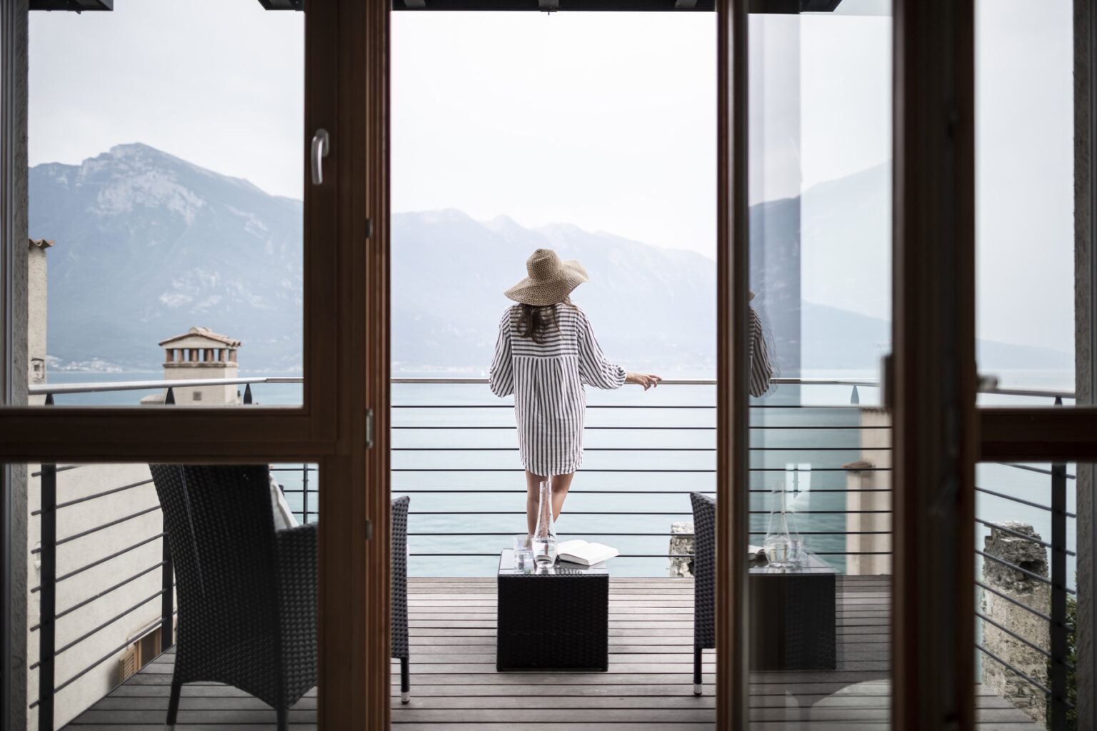 Woman in a white robe and hat standing on a balcony overlooking a serene lake and mountains, enjoying a peaceful retreat. | Only on SopranoVillas
