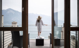 Woman in a white robe and hat standing on a balcony overlooking a serene lake and mountains, enjoying a peaceful retreat.
