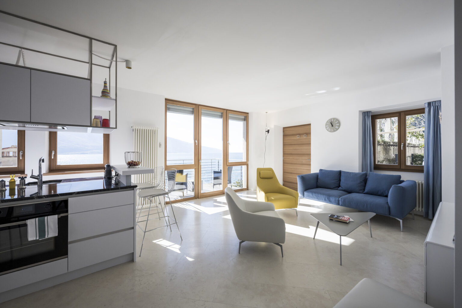 Modern living room with large windows, featuring a blue sofa, yellow accent chair, and minimalist kitchen with sleek cabinetry and appliances. Bright, airy space with natural light and a scenic view. | Only on SopranoVillas