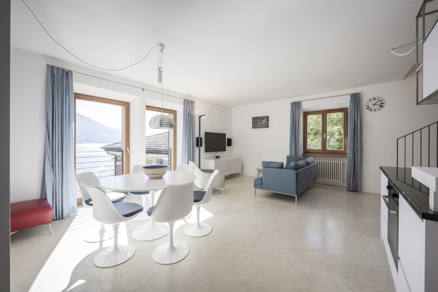 Modern living room and dining area with large windows showcasing a scenic view, featuring a round dining table with white chairs, a blue sofa, and minimalist decor. Natural light floods the space, highlighting the contemporary design and neutral color palette. | Only on SopranoVillas