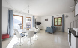 Modern living room and dining area with large windows showcasing a scenic view, featuring a round dining table with white chairs, a blue sofa, and minimalist decor. Natural light floods the space, highlighting the contemporary design and neutral color palette.