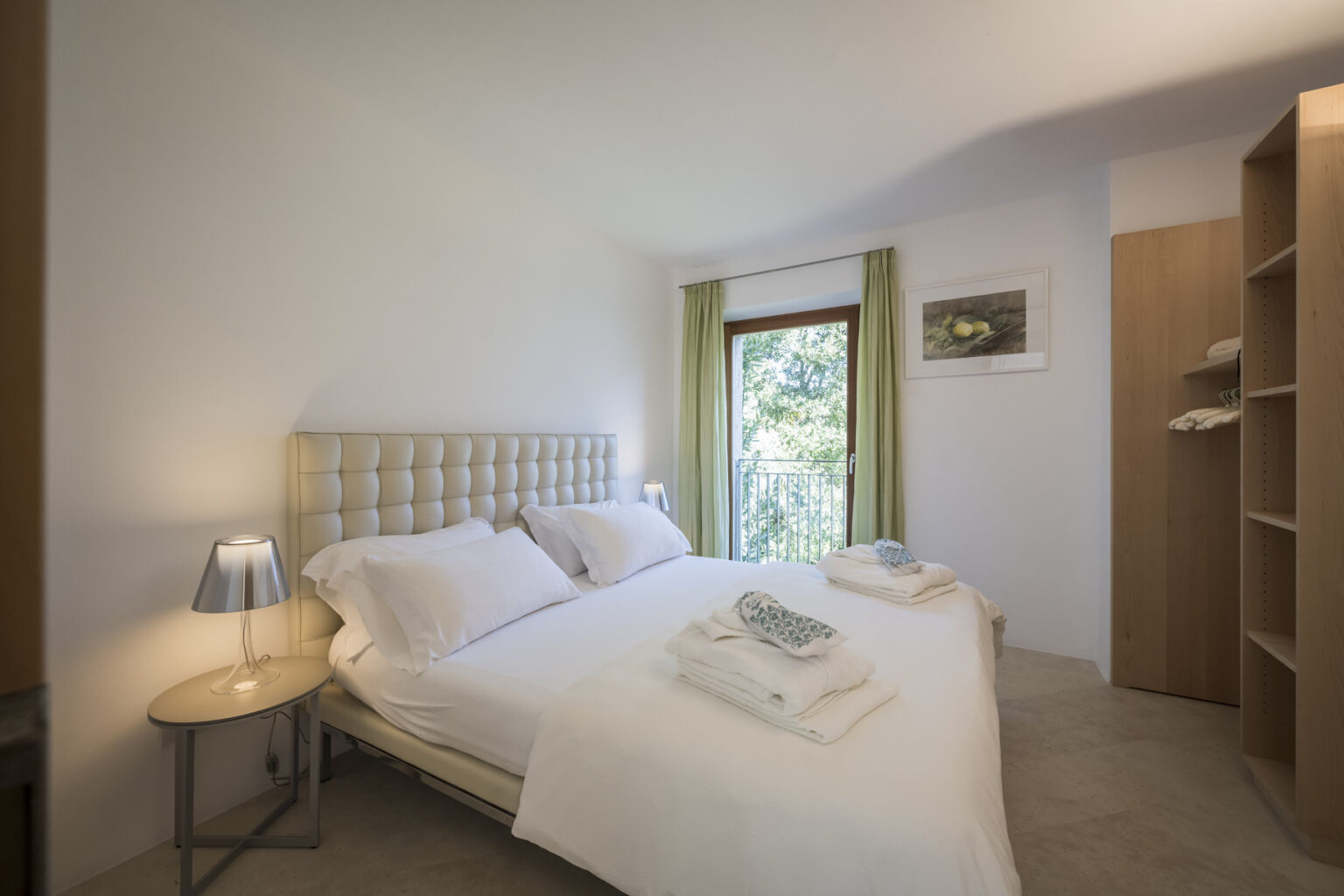 Cozy bedroom featuring a queen-sized bed with white linens, neatly arranged towels, a bedside table with a lamp, and large windows with green curtains, offering a bright and inviting atmosphere. | Only on SopranoVillas