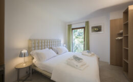 Cozy bedroom featuring a queen-sized bed with white linens, neatly arranged towels, a bedside table with a lamp, and large windows with green curtains, offering a bright and inviting atmosphere.