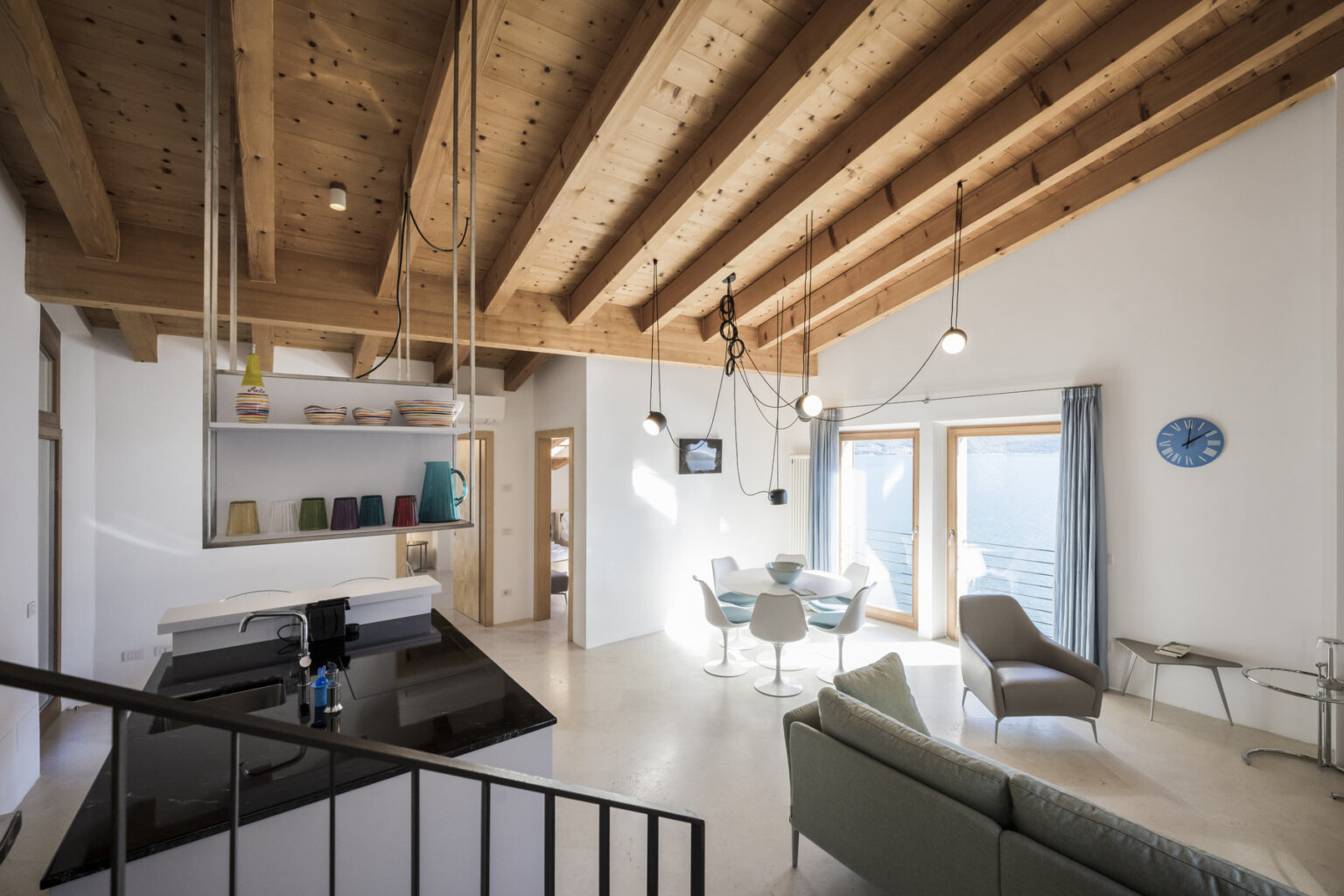 Modern interior design featuring a spacious living area with wooden beams, stylish furniture, and large windows that offer natural light. The open layout includes a dining table, a sleek kitchen with black countertops, and contemporary decor elements, creating a warm and inviting atmosphere. | Only on SopranoVillas
