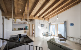 Modern interior design featuring a spacious living area with wooden beams, stylish furniture, and large windows that offer natural light. The open layout includes a dining table, a sleek kitchen with black countertops, and contemporary decor elements, creating a warm and inviting atmosphere.
