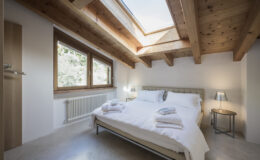 Modern bedroom with wooden beams, large windows, and a cozy double bed adorned with fresh towels, creating a bright and inviting atmosphere.