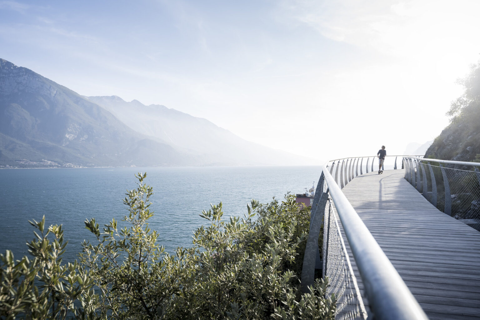 Scenic view of a wooden walkway along a serene lake, surrounded by mountains and greenery, with a person walking towards the horizon under a bright sky. | Only on SopranoVillas