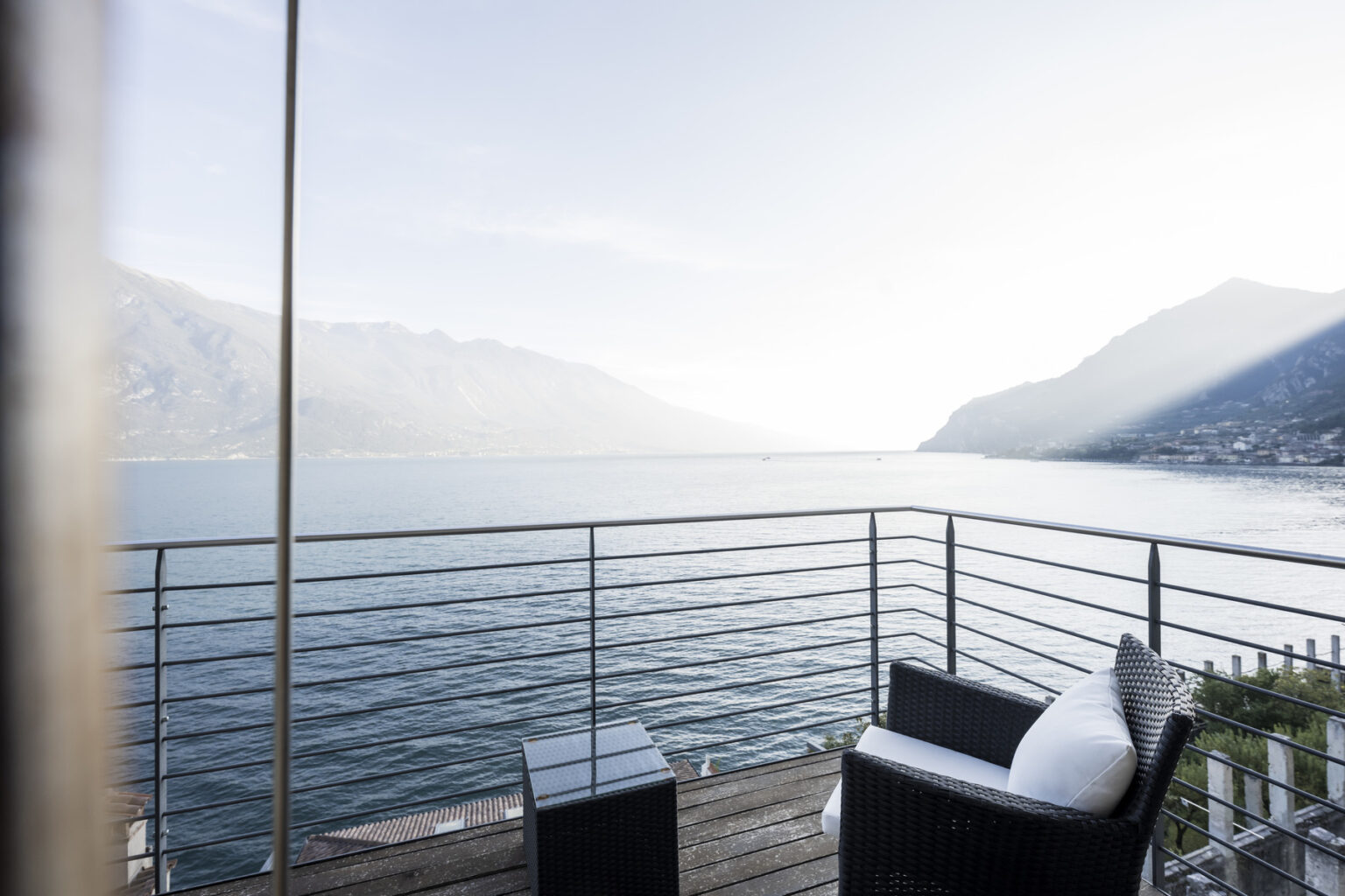 Modern balcony overlooking a serene lake surrounded by mountains, featuring stylish outdoor furniture and a peaceful atmosphere. | Only on SopranoVillas