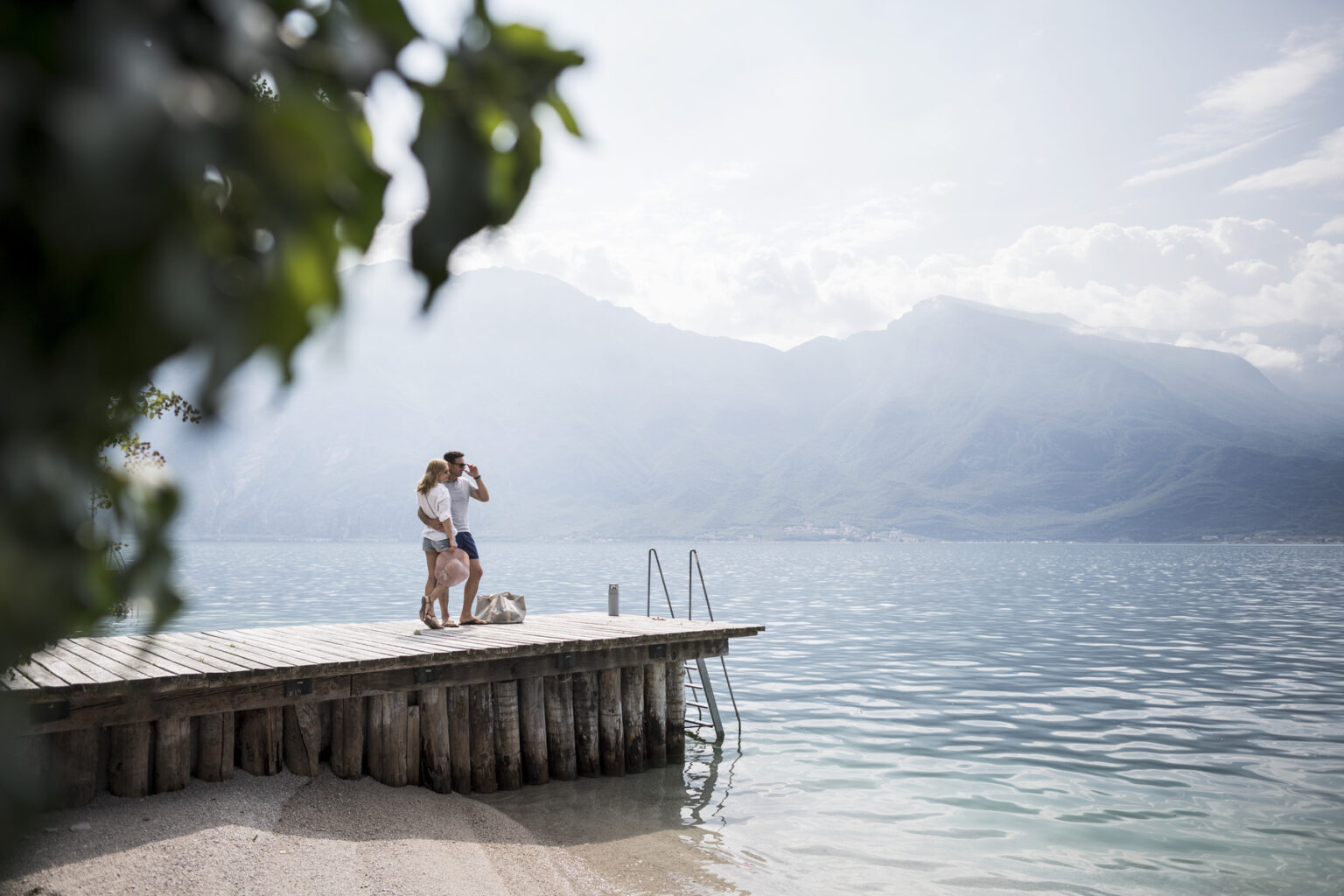 Couple enjoying a romantic moment on a wooden dock by a serene lake, with mountains in the background and a cloudy sky. | Only on SopranoVillas