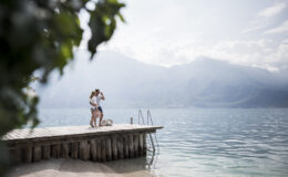 Couple enjoying a romantic moment on a wooden dock by a serene lake, with mountains in the background and a cloudy sky.