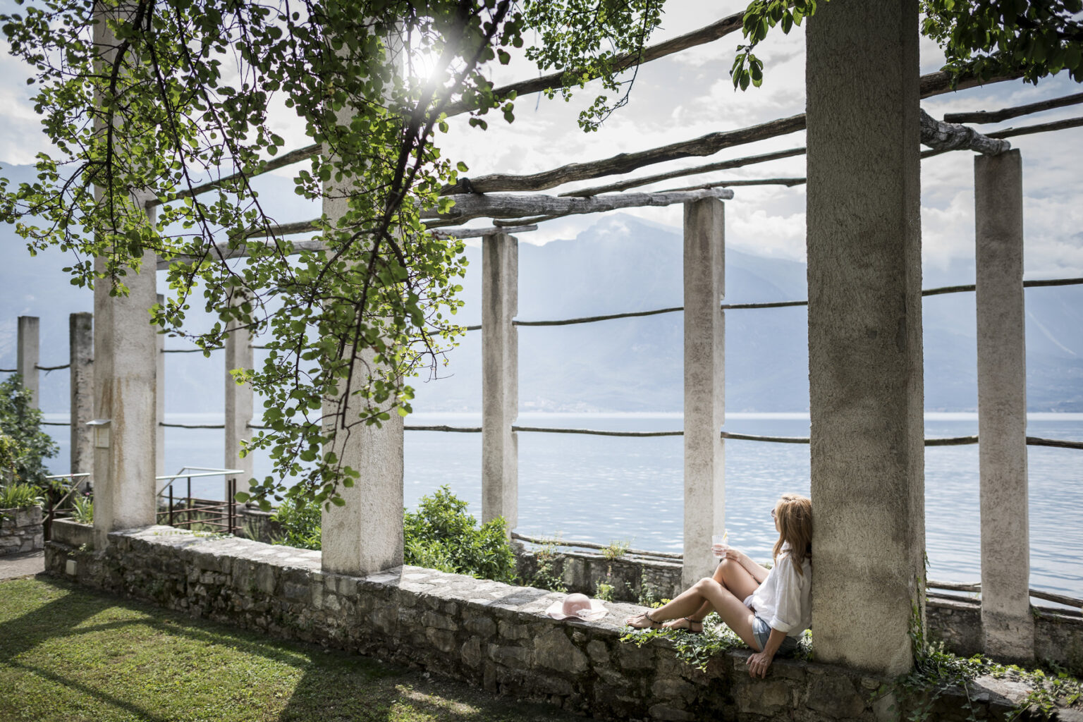 A young woman sitting on a stone wall under a leafy canopy, enjoying a serene view of a lake surrounded by mountains, with sunlight filtering through the columns of an open structure. | Only on SopranoVillas