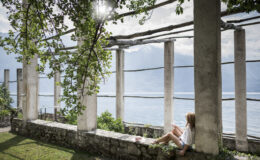 A young woman sitting on a stone wall under a leafy canopy, enjoying a serene view of a lake surrounded by mountains, with sunlight filtering through the columns of an open structure.