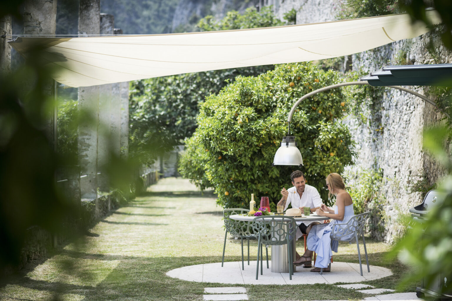 Couple enjoying a meal together at a stylish outdoor dining table under a shaded canopy, surrounded by lush greenery in a serene garden setting. | Only on SopranoVillas