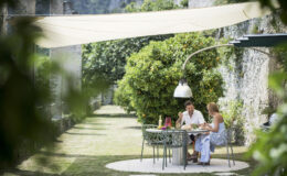 Couple enjoying a meal together at a stylish outdoor dining table under a shaded canopy, surrounded by lush greenery in a serene garden setting.