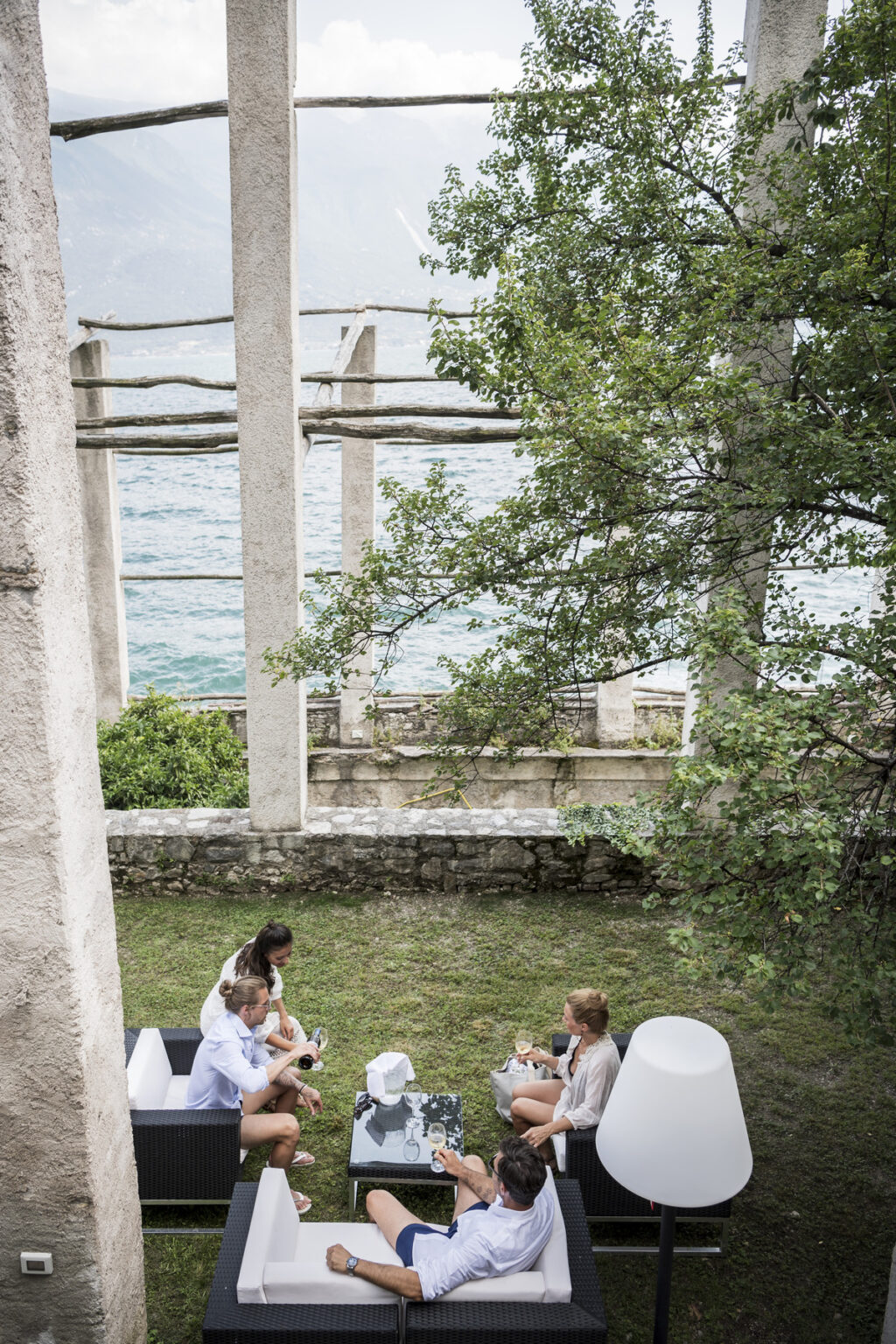 A person sitting on the grass near the ruins of a structure by the water, surrounded by trees and greenery, with a serene view of the lake in the background. | Only on SopranoVillas