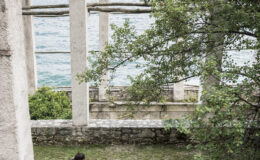 A person sitting on the grass near the ruins of a structure by the water, surrounded by trees and greenery, with a serene view of the lake in the background.
