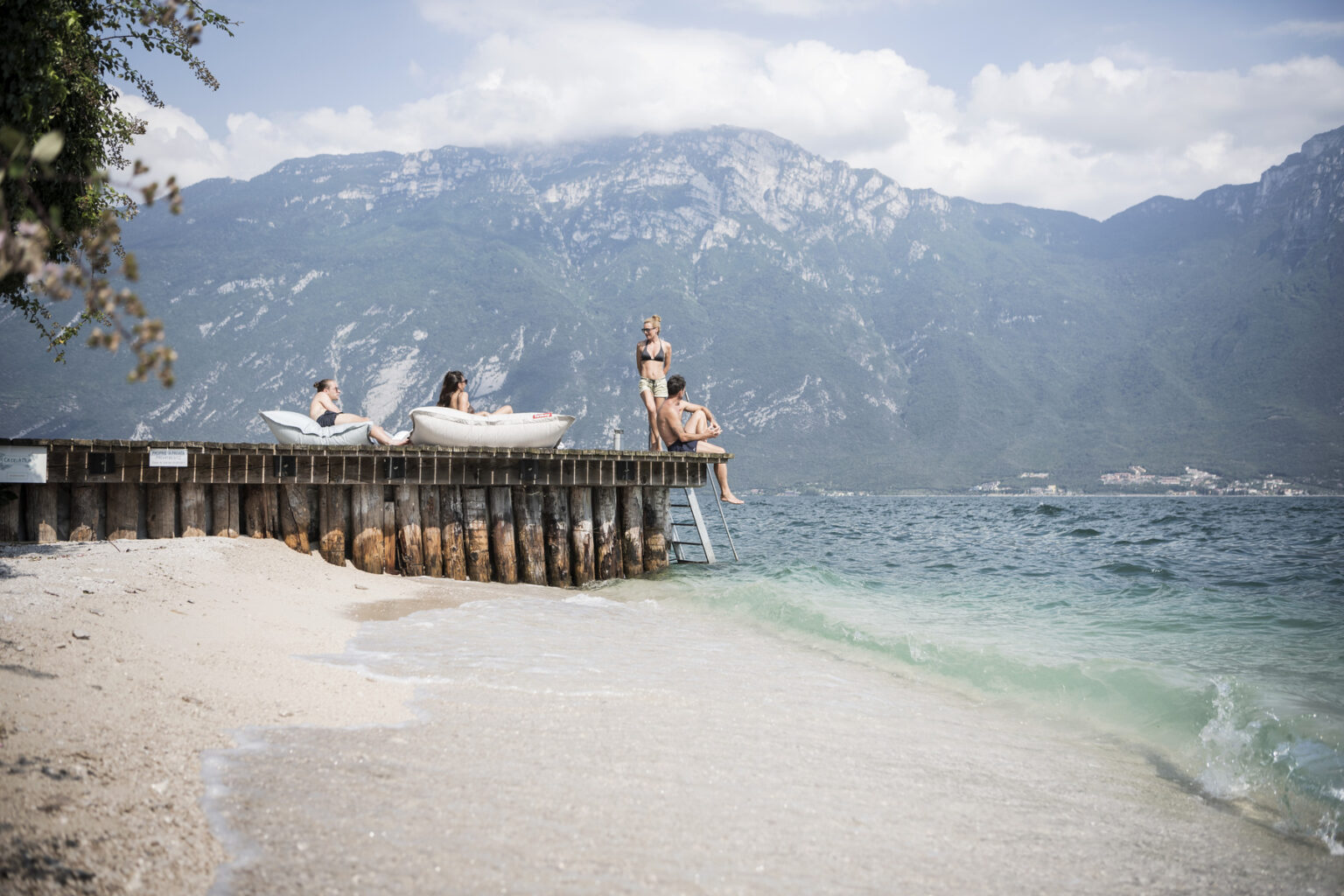People relaxing on a wooden pier by a lake, with mountains in the background and sandy beach in the foreground, capturing a serene summer day. | Only on SopranoVillas