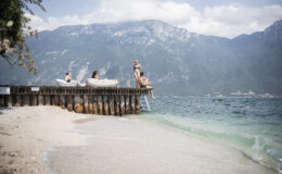 People relaxing on a wooden pier by a lake, with mountains in the background and sandy beach in the foreground, capturing a serene summer day.