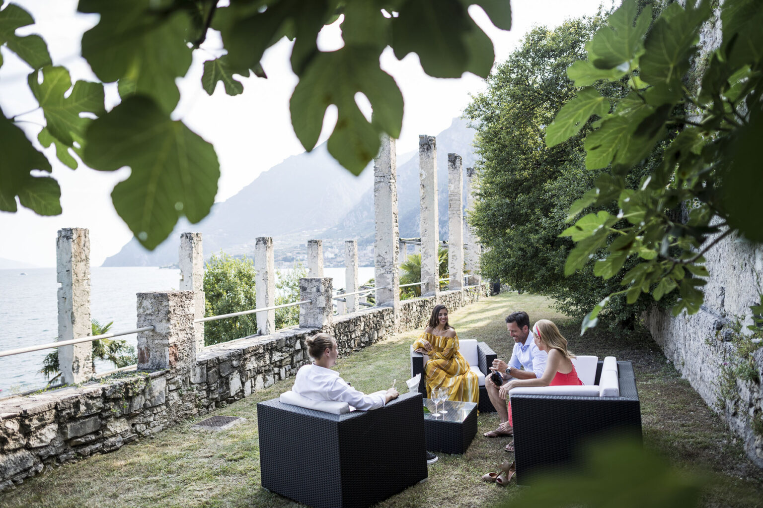 Group of four people enjoying a conversation outdoors by a lake, seated on modern black furniture surrounded by greenery and historic stone ruins. | Only on SopranoVillas