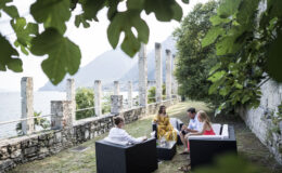 Group of four people enjoying a conversation outdoors by a lake, seated on modern black furniture surrounded by greenery and historic stone ruins.